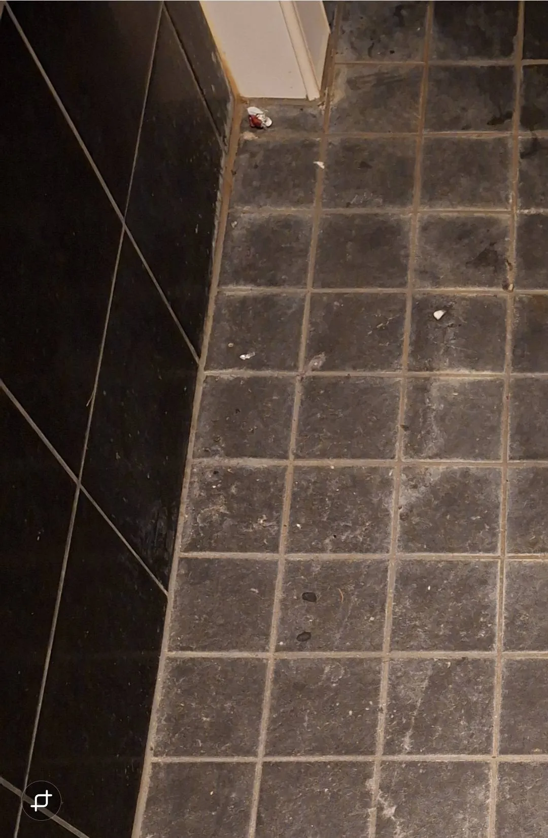 Lattianäkymä, jossa on tummat laatat, valkoiset saumat ja roskia, sekä osa mustasta seinästä ja valkoisesta jalkalistasta.