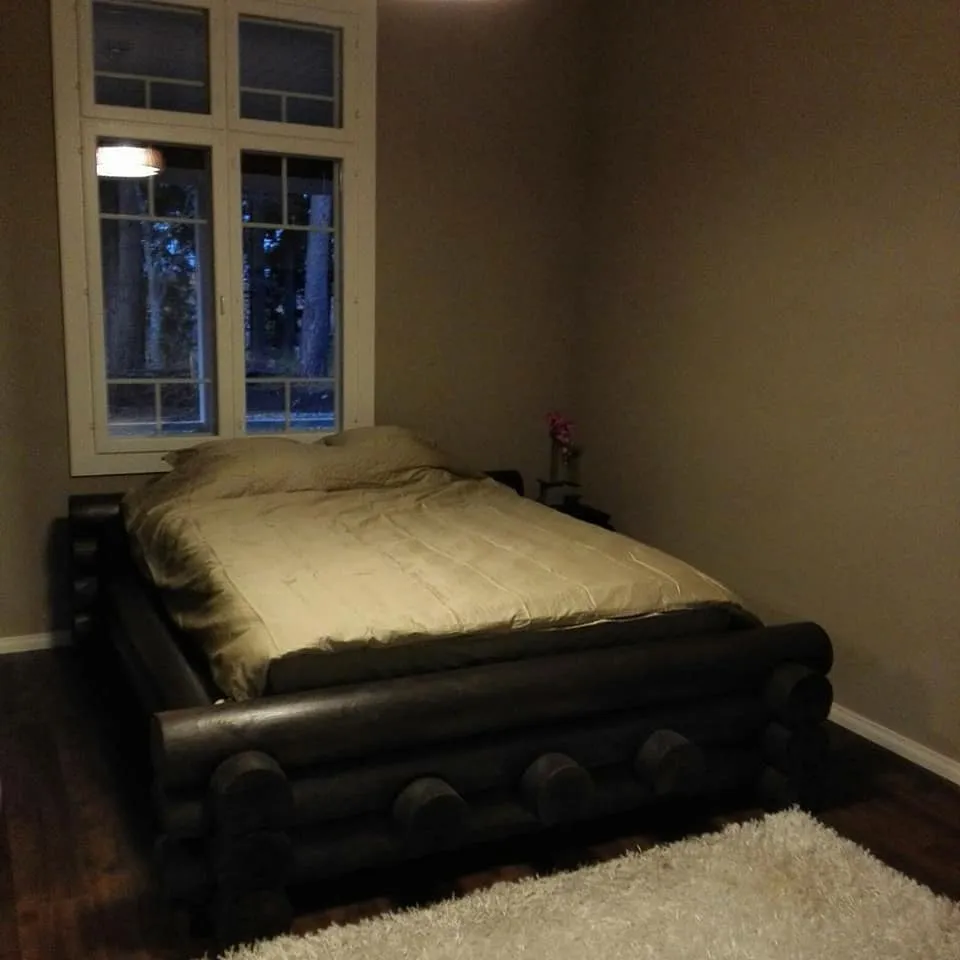 Makuuhuoneessa on hirsirunkoinen sänky, jonka päällä on beige peitto ja tyynyt. Ikkunasta näkyy metsää, ja lattialla on valkoinen matto.