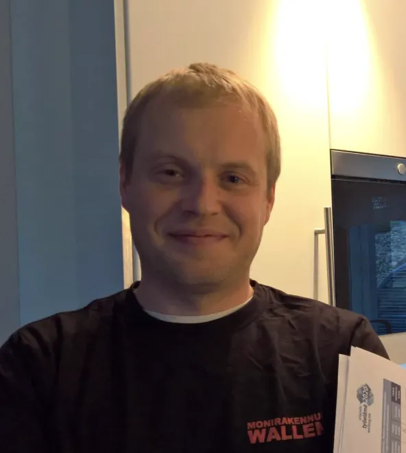 Lähikuva hymyilevästä vaaleahiuksisesta miehestä, jolla on musta paita, jossa on punainen teksti "MONIRAKENNU WALLEN", ja hän pitelee papereita keittiössä, jossa on valkoiset kaapit ja teräksinen uuni.