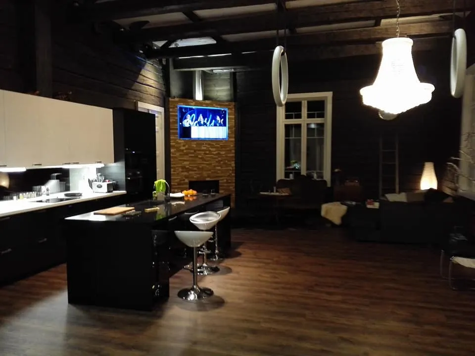 Moderni keittiö ja olohuone, jossa on mustavalkoiset kaapit, saareke, TV, kattokruunu ja puulattia.