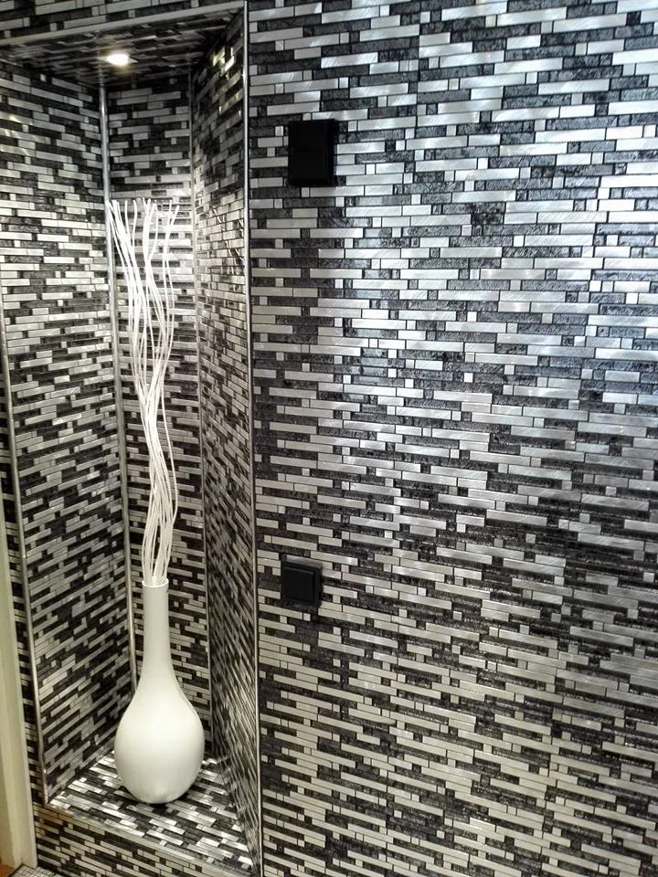 Sisustuskuva mosaiikkiseinästä, jossa valkoinen maljakko ja koristeelliset oksat syvennyksessä, sekä kaksi mustaa pistorasiaa seinällä.