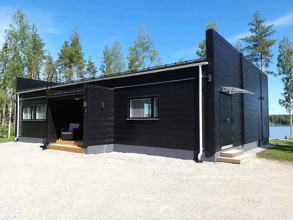 Moderni, musta puurakennus valkoisilla yksityiskohdilla, sijaitsee aurinkoisessa ympäristössä, jossa on puita ja järvi taustalla.