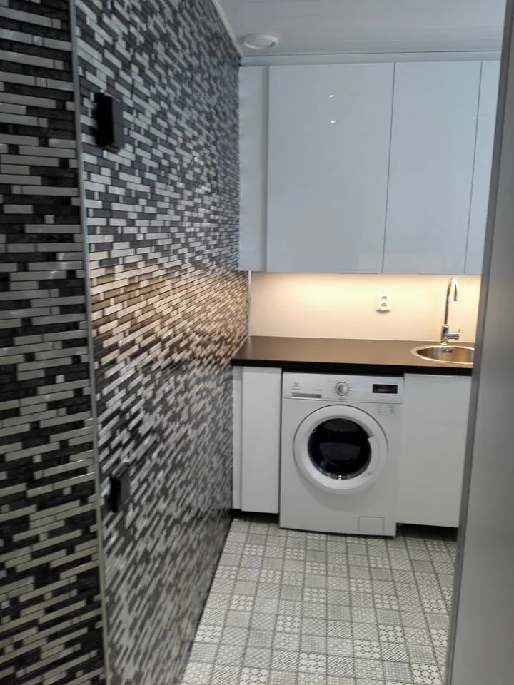 Pieni pesuhuone, jossa on mosaiikkiseinä, valkoiset kaapit, pesukone ja harmaa lattia.