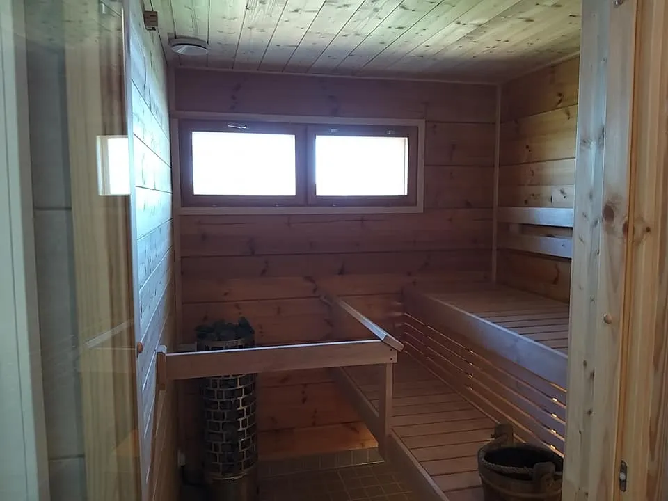 Puupaneloitu sauna, jossa on kaksi ikkunaa, penkit ja kiuas. Valoisa ja lämmin tunnelma.