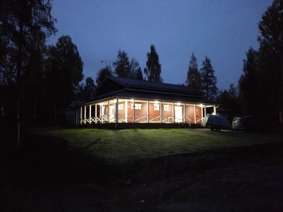 Hämärässä kuvassa näkyy punainen talo, jonka edessä on valaistu nurmikko ja taustalla tumma metsä.