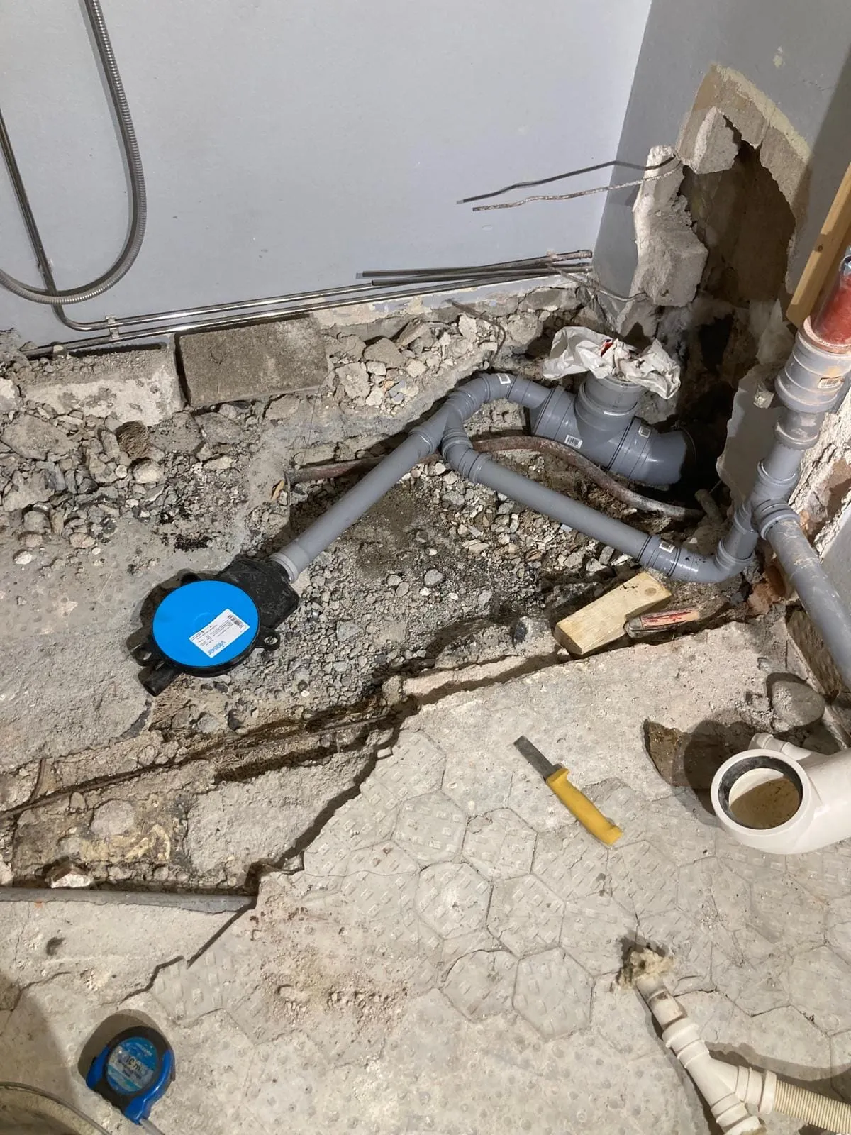 Lattialla on harmaita muoviputkia, sininen kansi, keltainen ruuvimeisseli ja mittanauha, valaistus on kirkas.