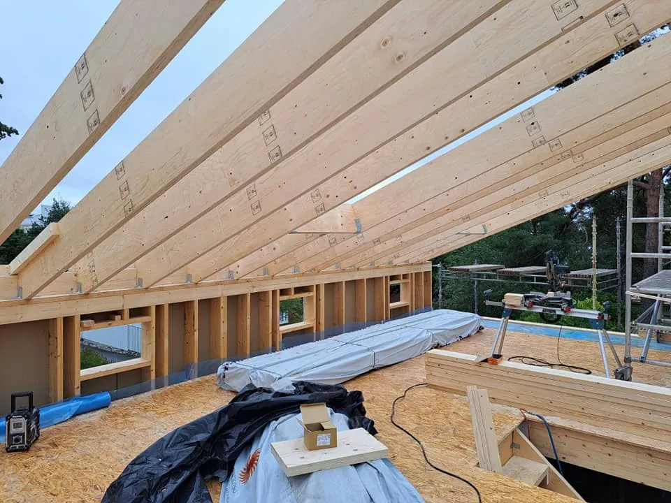 Rakenteilla oleva puurakenteinen talo, jossa näkyy kattotuolit, seinät ja rakennustarvikkeita. Valoisa ja avoin tila.