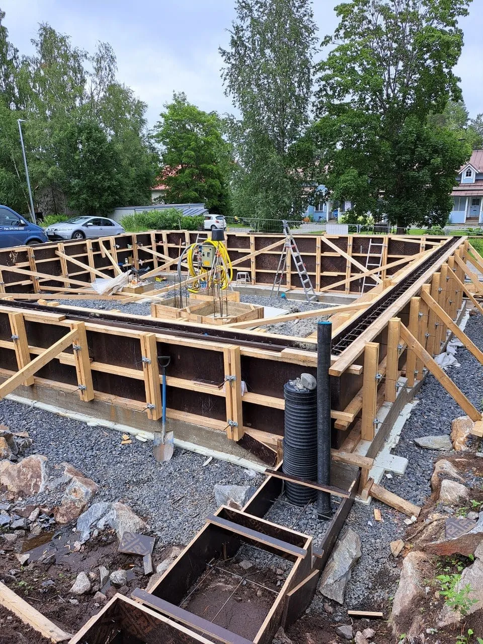 Rakennustyömaa, jossa on puinen muotti tulevaa betonirakennetta varten, harmaa kivituhka ja vihreitä puita taustalla, valoisa sää.