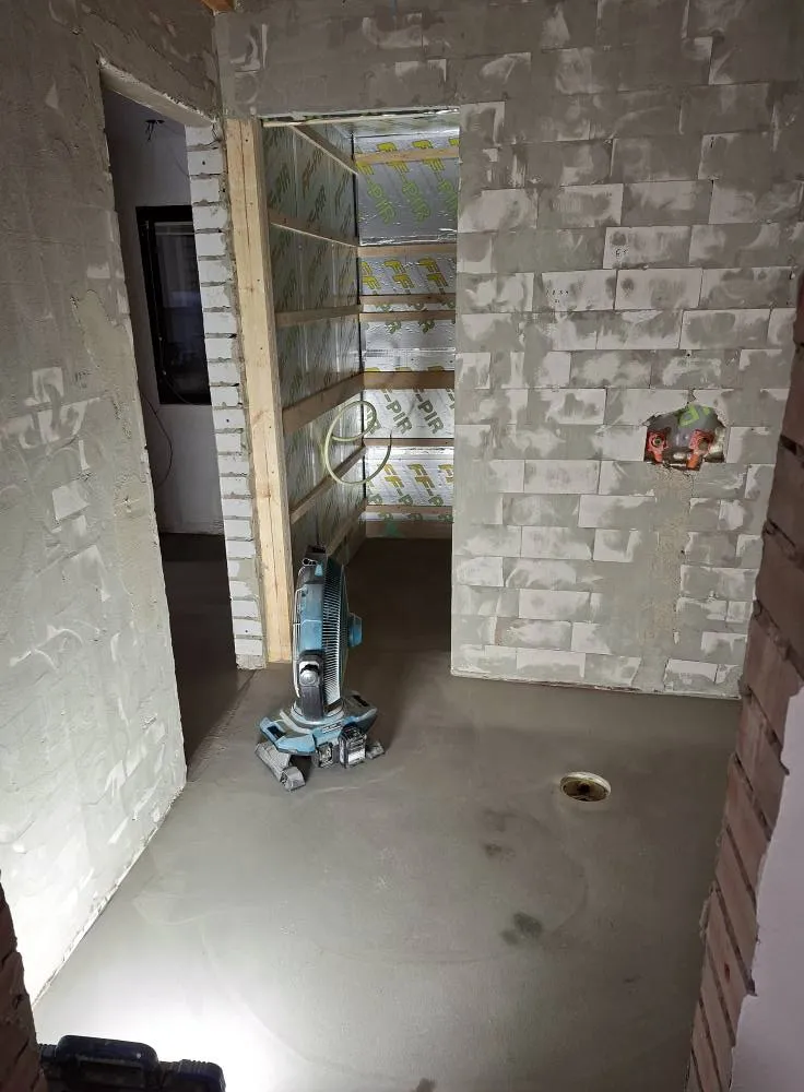 Keskeneräinen huone, jossa on sementtilattia, tuuletin ja paljaat seinät, valaistus on kirkas.