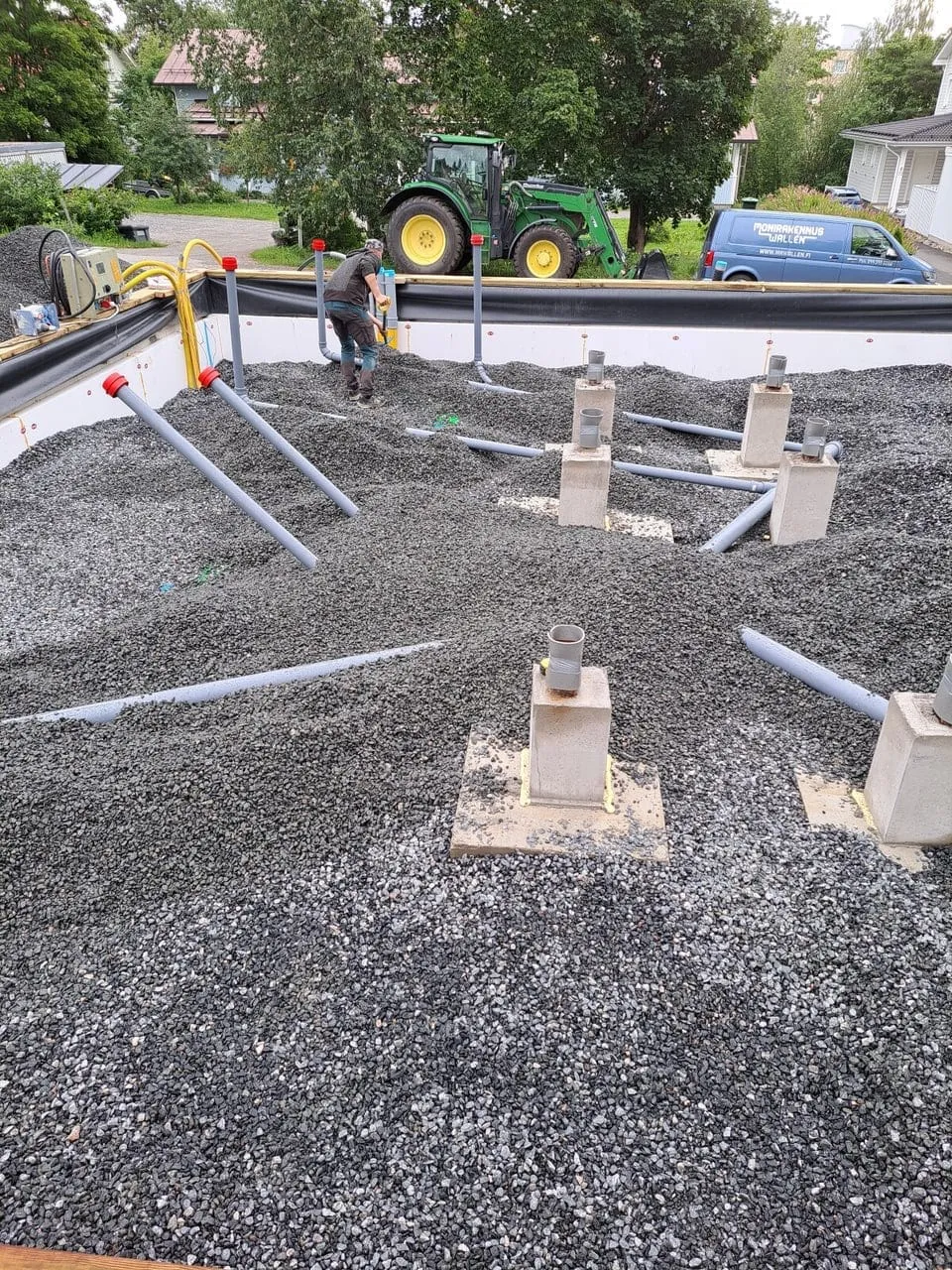 Rakennustyömaa, jossa on harmaata soraa, putkia ja betonipylväitä. Traktori ja pakettiauto taustalla. Kirkas päivänvalo.