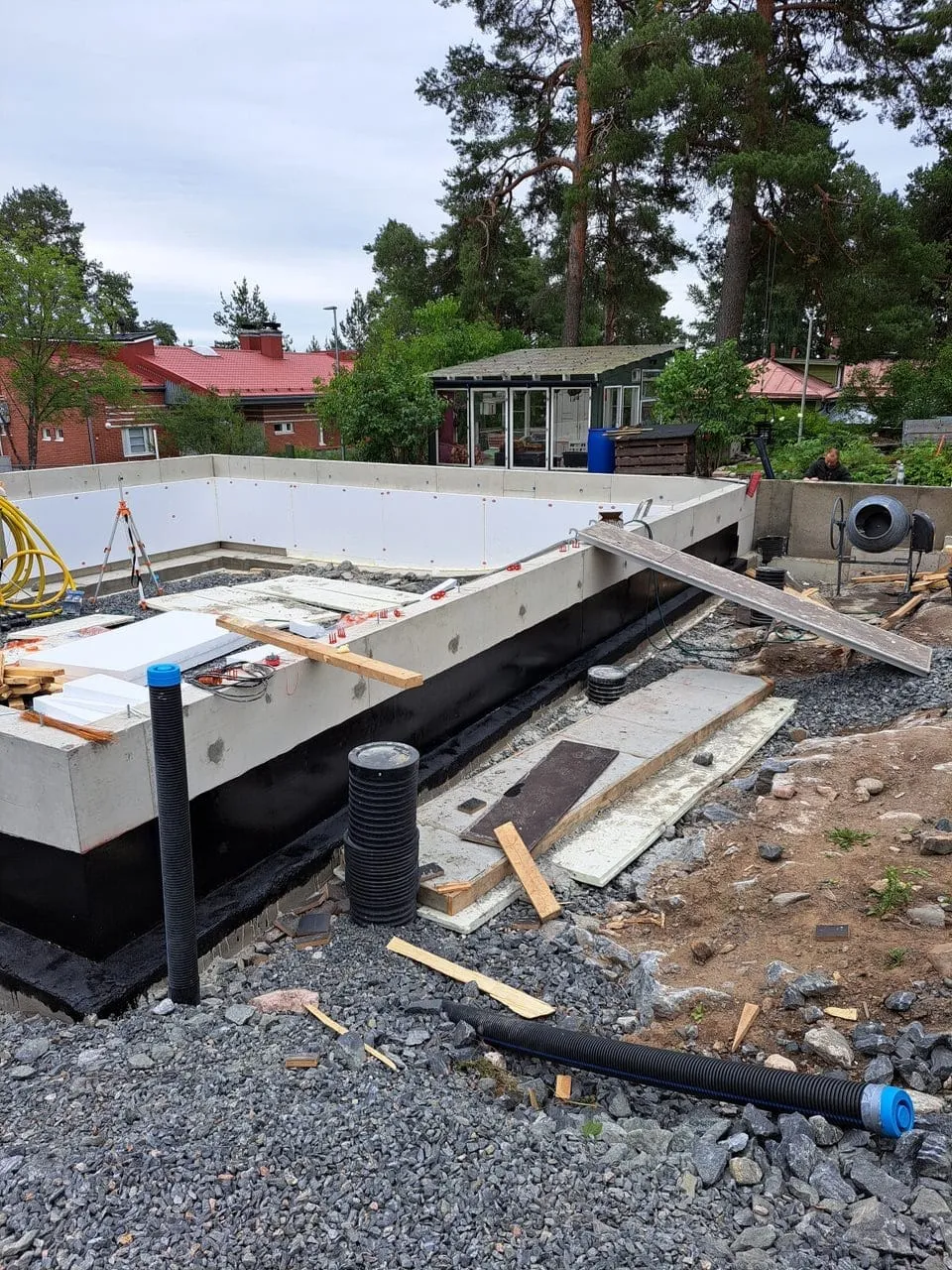 Rakennustyömaa ulkona, jossa näkyy betoniperustus, musta vedeneristys, harmaa sora ja puita taustalla, valaistus on luonnollinen ja pilvinen.