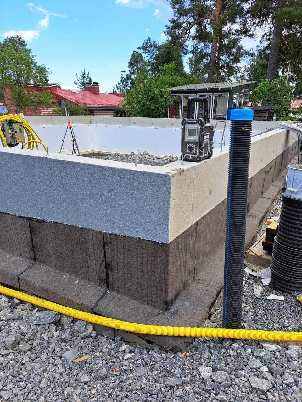 Ulkona on rakennustyömaa, jossa näkyy betoninen perustus, keltainen putki ja Makita-radio. Valoisa sää.