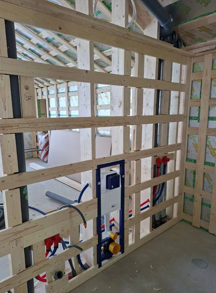 Rakenteilla oleva kylpyhuone, jossa näkyy puurunkoinen seinä, putkistoja ja Geberit-seinä-WC:n runko. Valoisa ja keskeneräinen tila.