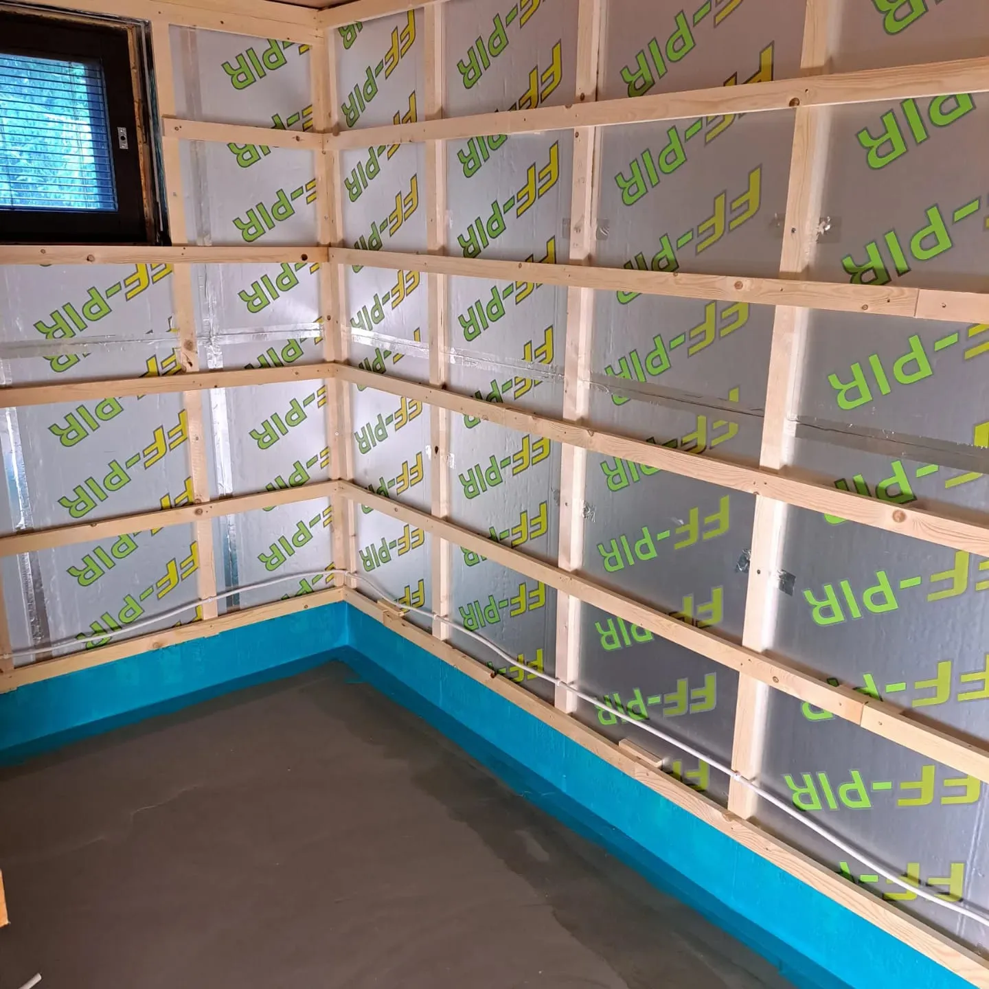 Keskeneräinen huone, jossa on puurunkoinen seinä, eristyslevyt, sininen kosteussulku ja betonialattia, valaistus on kirkas.