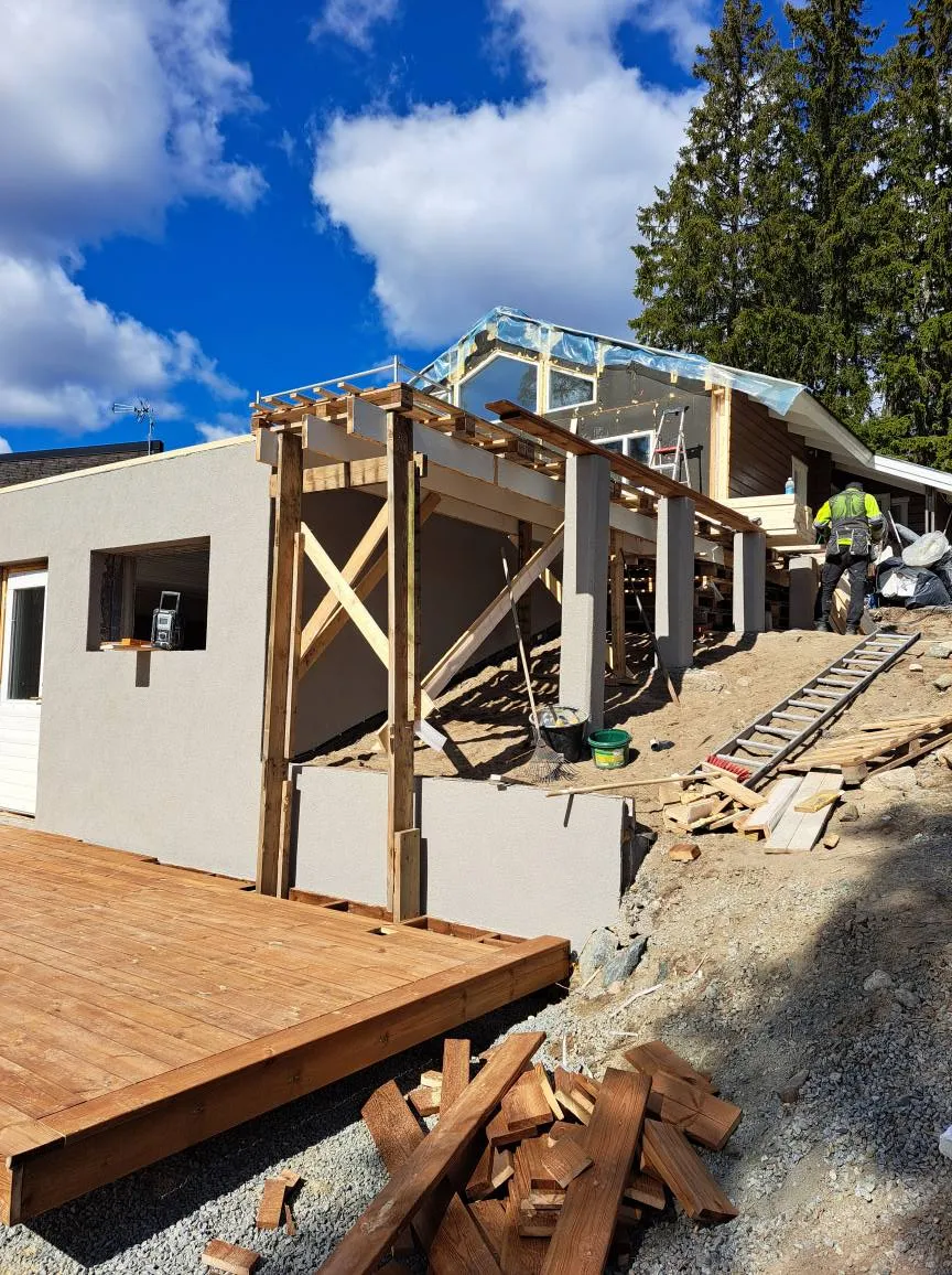 Rakenteilla oleva talo aurinkoisena päivänä, jossa näkyy puinen terassi, harmaa seinä, puurakenteet ja taustalla puita sekä sininen taivas pilvillä.