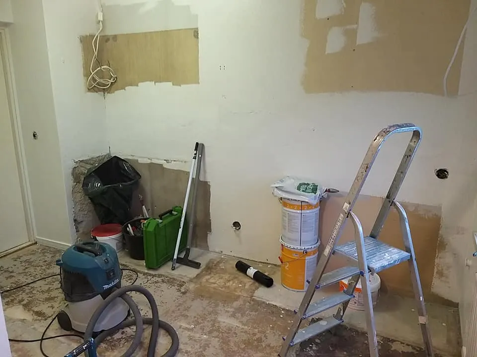 Huoneessa on remontti meneillään, jossa näkyy maalipurkkeja, tikkaat, imuri ja muita remonttitarvikkeita. Seinät ovat osittain maalattuja ja osittain paljaita.