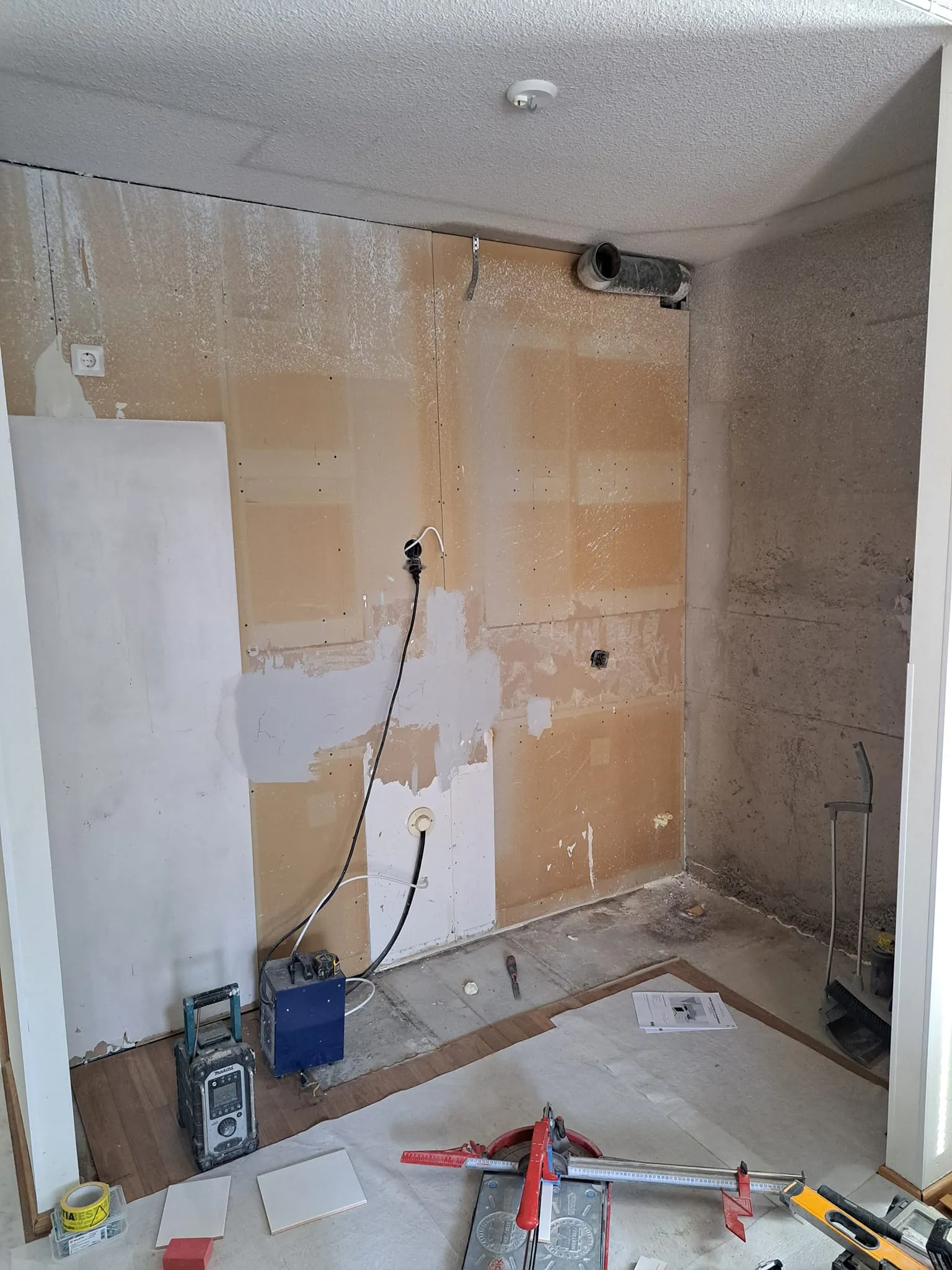 Huone remontin alla, jossa näkyy työkaluja, laattoja ja sähkölaitteita. Seinät ovat osittain peitetty levyillä ja lattia on suojattu paperilla.