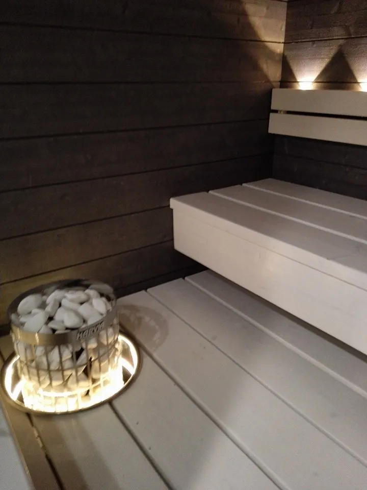 Saunan sisustus, jossa on valaistu kivikori ja valkoiset laudat.