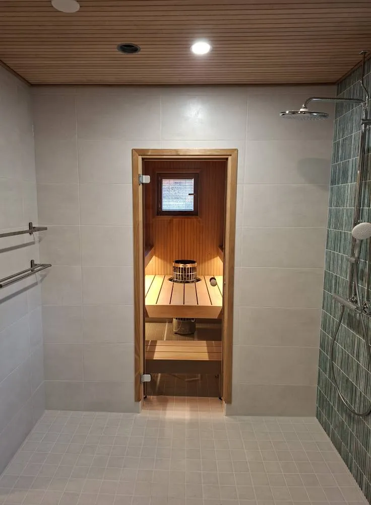 Kylpyhuoneessa on sauna, jossa on puinen sisustus ja lasiovi. Suihku on oikealla ja pyyheteline vasemmalla. Lattialaatat ovat vaaleat ja katto on puupaneloitu.