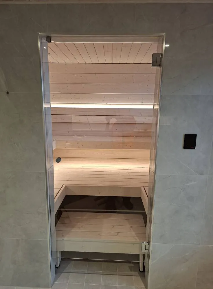 Saunan lasiovi, vaaleat puupaneelit, himmeä valaistus ja harmaat laatat.