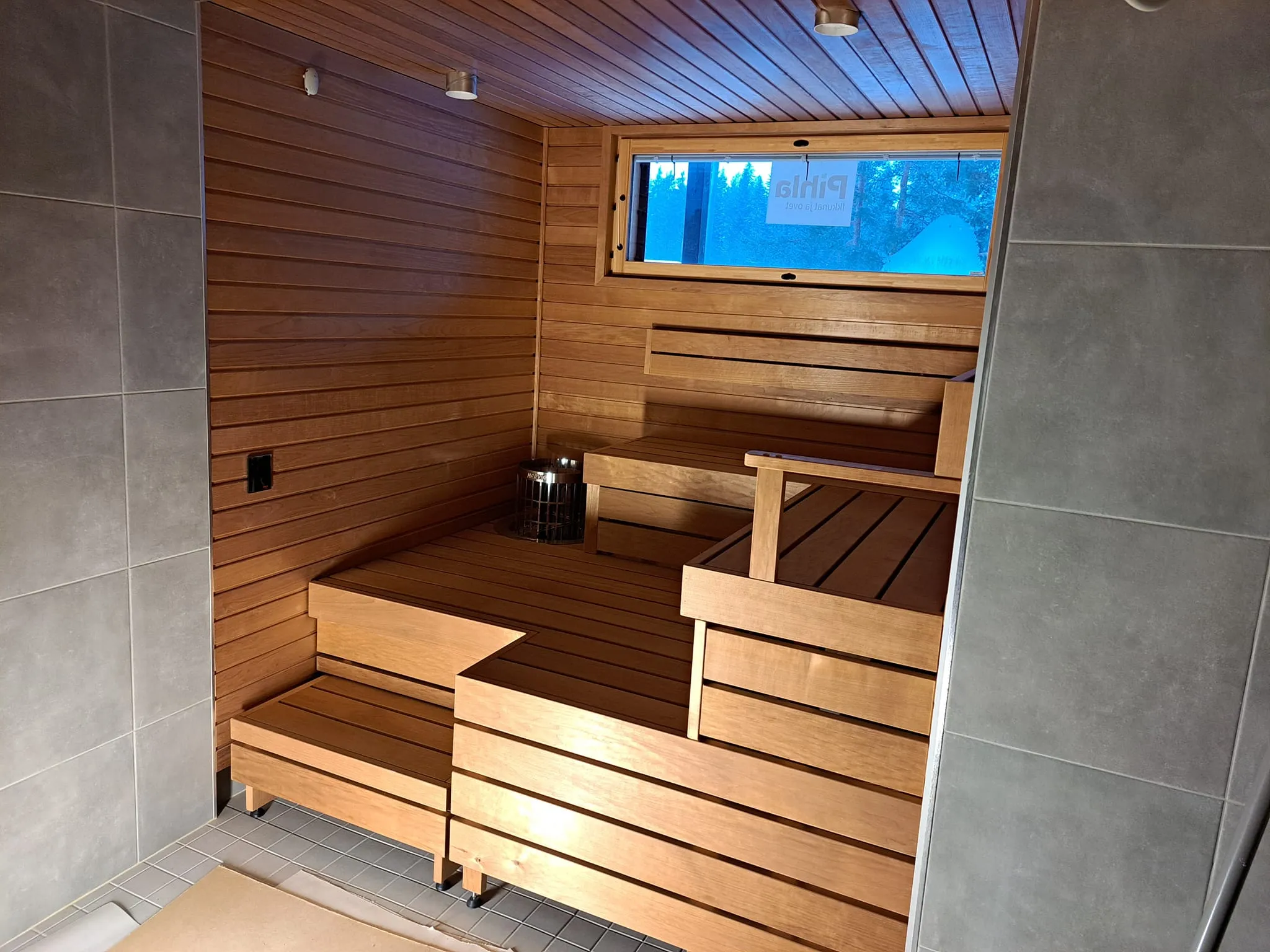 Saunan sisätila puupaneloinnilla, lauteilla ja pienellä ikkunalla, jossa näkyy ulkona oleva metsä.