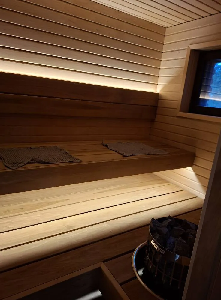 Sauna, jossa on puupanelointi, lauteet, kaksi pyyhettä ja kiuas, jossa on mustia kiviä. Valo tulee lauteiden alta ja pienestä ikkunasta.