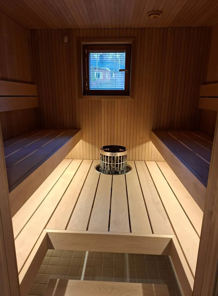 Sauna, jossa on puupaneelit, penkit ja kiuas, jossa on kiviä metallisessa korissa. Ikkunasta näkyy ulos.