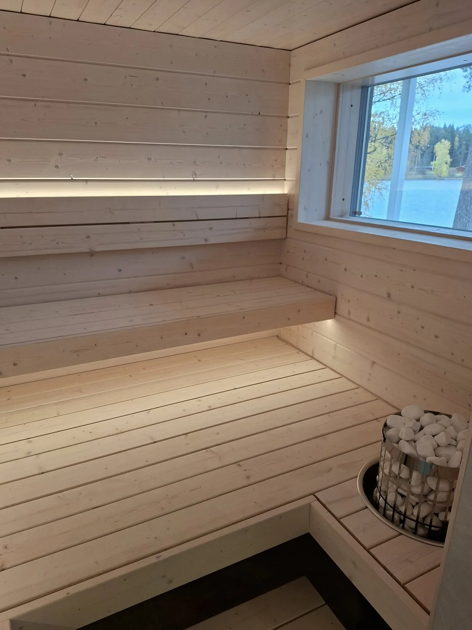 Vaalea puusauna, jossa on ikkuna, lauteet ja kiuas kivillä. Tunnelmallinen valaistus.