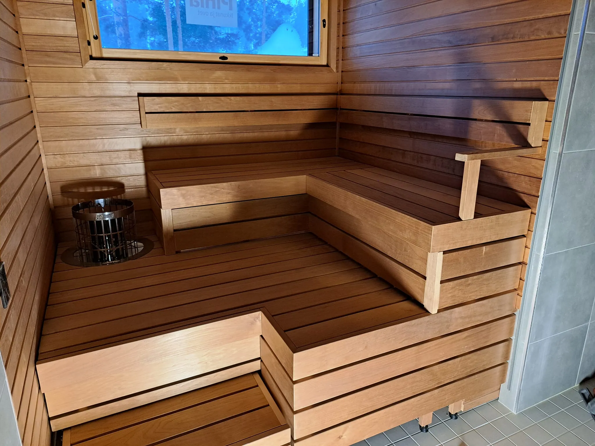 Sauna, jossa on puupanelointi, penkit ja kiuas, sekä ikkuna, josta näkyy ulos.