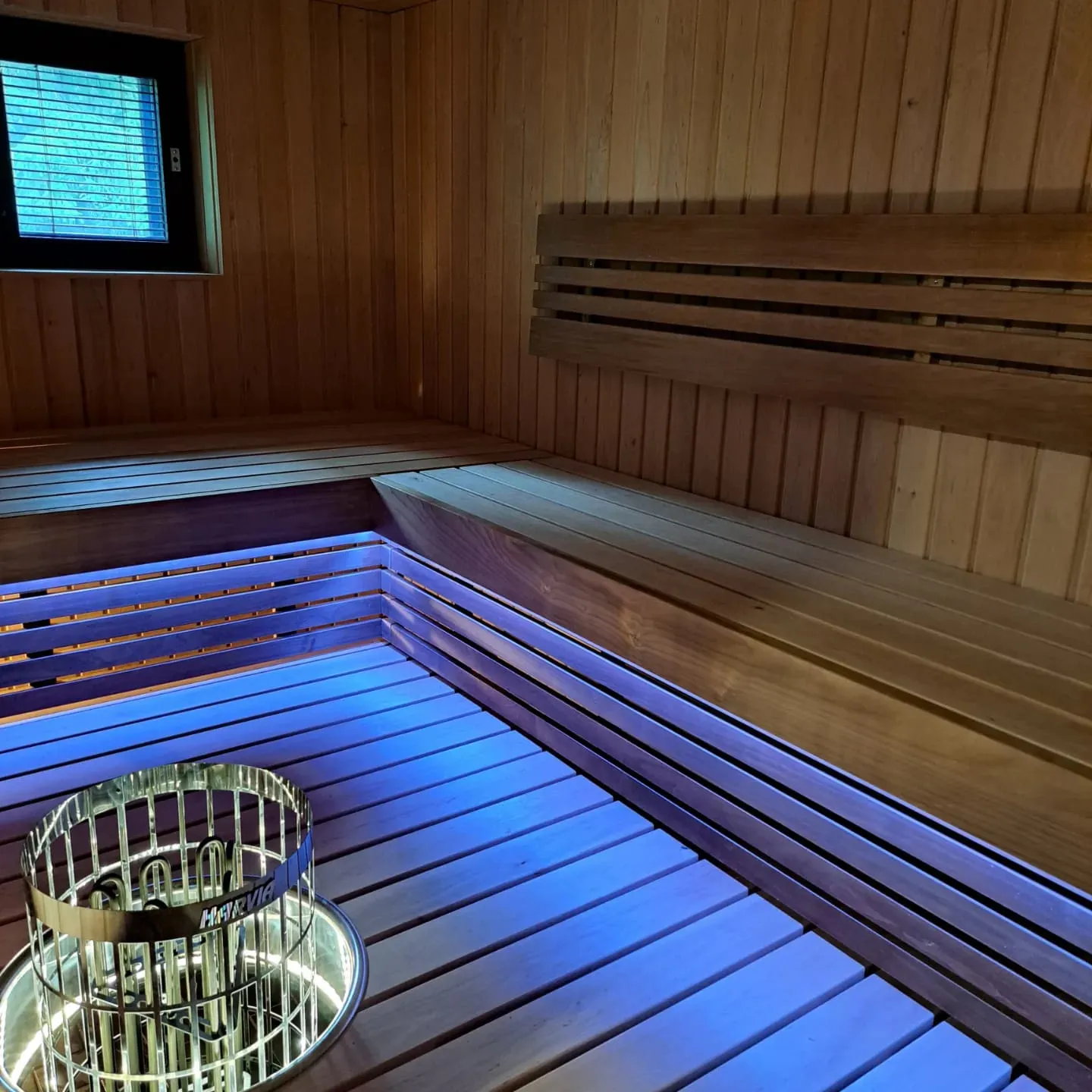 Puinen sauna, jossa on penkit, ikkuna ja Harvia-kiuas, valaistu sinisellä valolla.