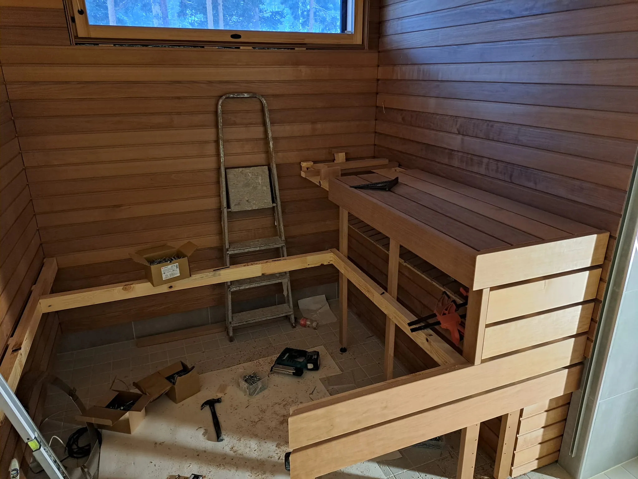 Kuva esittää saunan sisätiloja rakennusvaiheessa, jossa näkyy puupanelointi, ikkuna, tikkaat ja osittain rakennettu lauteet. Valoisa ja puun sävyinen.