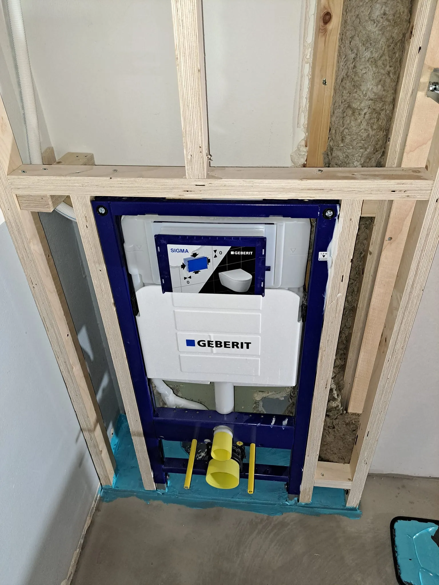 Kuva näyttää Geberit-seinä-WC:n asennuksen puurungossa, jossa on eristys ja sininen vesieristys lattialla.