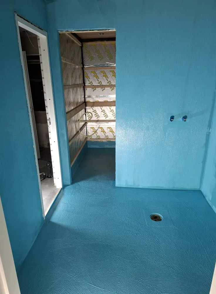 Sininen huone, jossa on avoin ovi ja viereinen tila, jossa on eristys ja puu. Lattia ja seinät on maalattu sinisellä, ja huoneessa on viemäri.