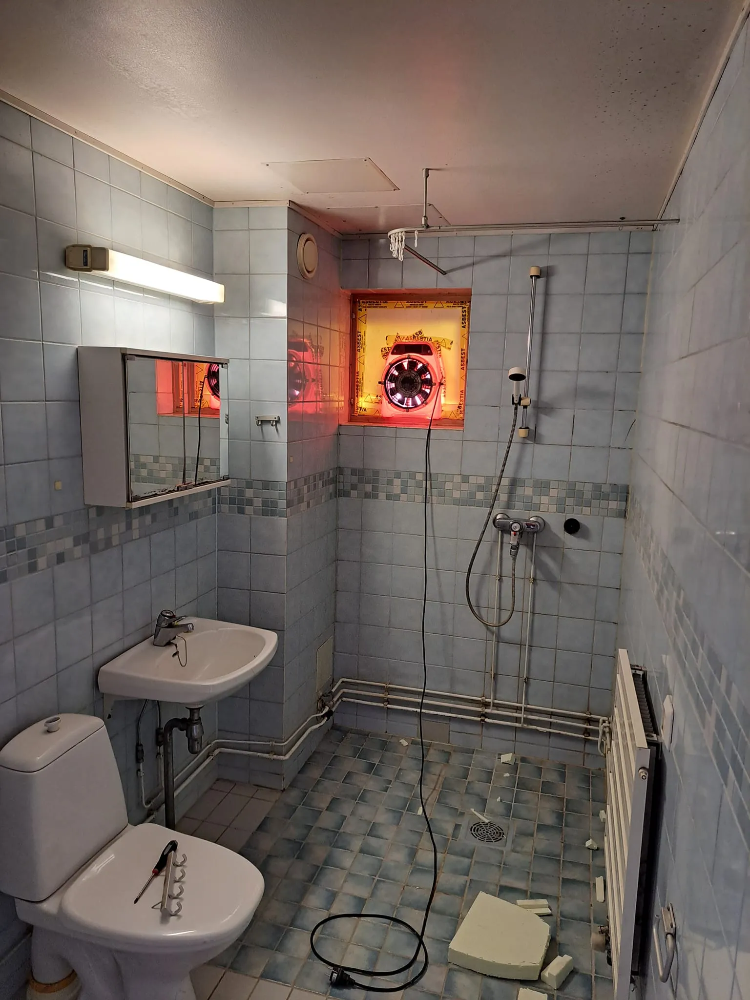 Kylpyhuoneessa on siniset laatat, valkoinen pesuallas ja wc-istuin, jonka päällä on työkaluja. Ikkunassa on punainen valo ja lattialla on valkoisia paloja.