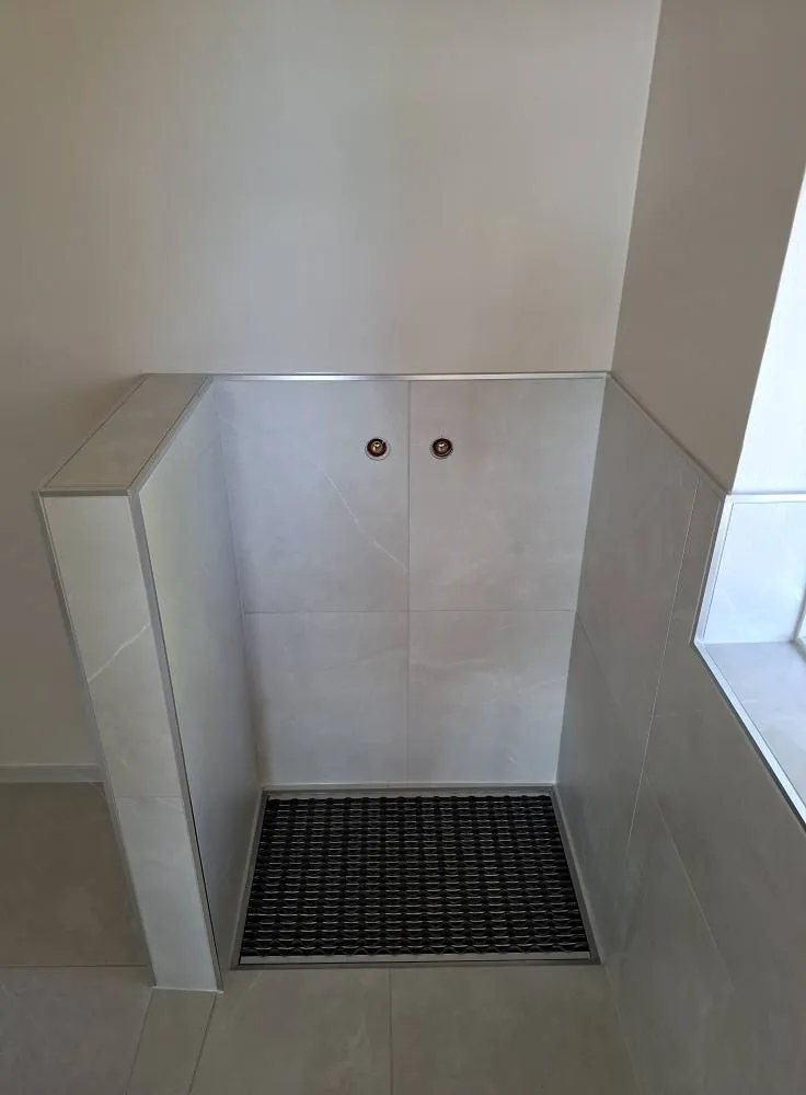 Laatoitettu suihkunurkkaus, jossa on musta ritilämatto, kaksi pyöreää reikää seinässä ja harmaa laatoitus.