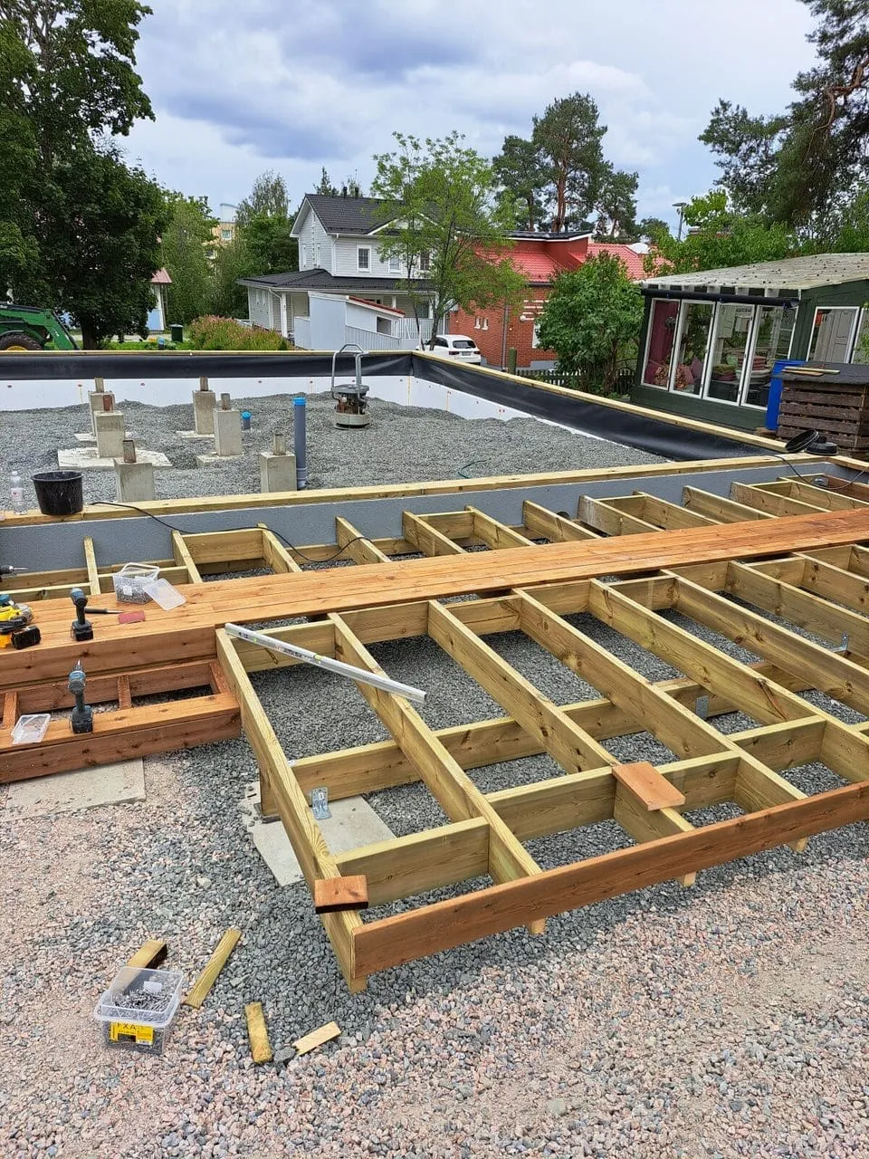 Rakenteilla oleva puinen terassi kivipohjalla, taustalla taloja ja vihreää kasvillisuutta, päivänvalo.