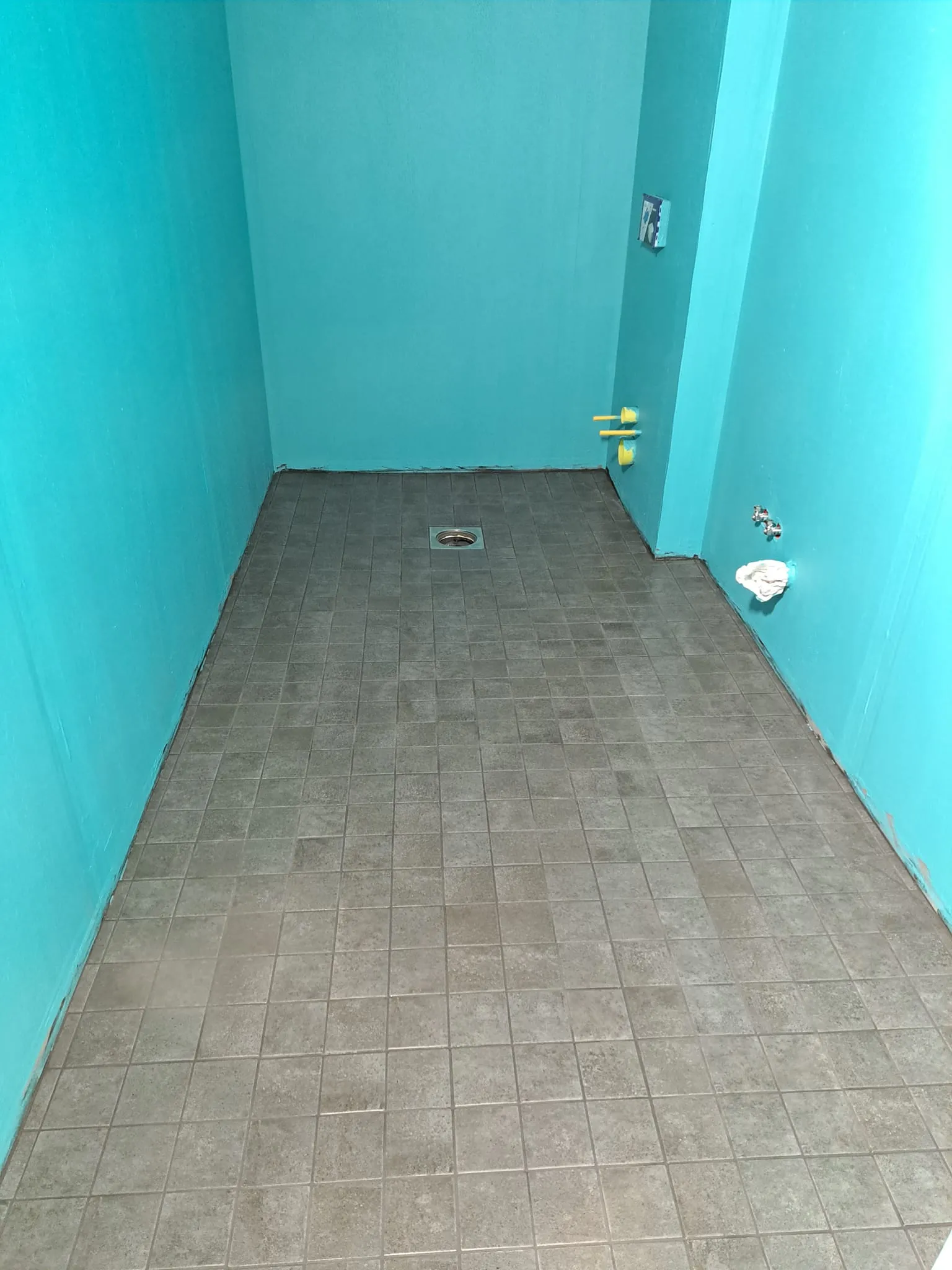 Turkoosiseinäinen huone, jossa on harmaat laatat, lattiakaivo, putket ja vesihana.