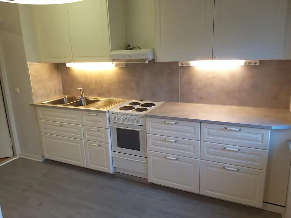 Vaaleassa keittiössä on valkoiset kaapit, harmaa työtaso ja harmaa taustalevy. Keittiössä on myös liesi ja pesuallas.