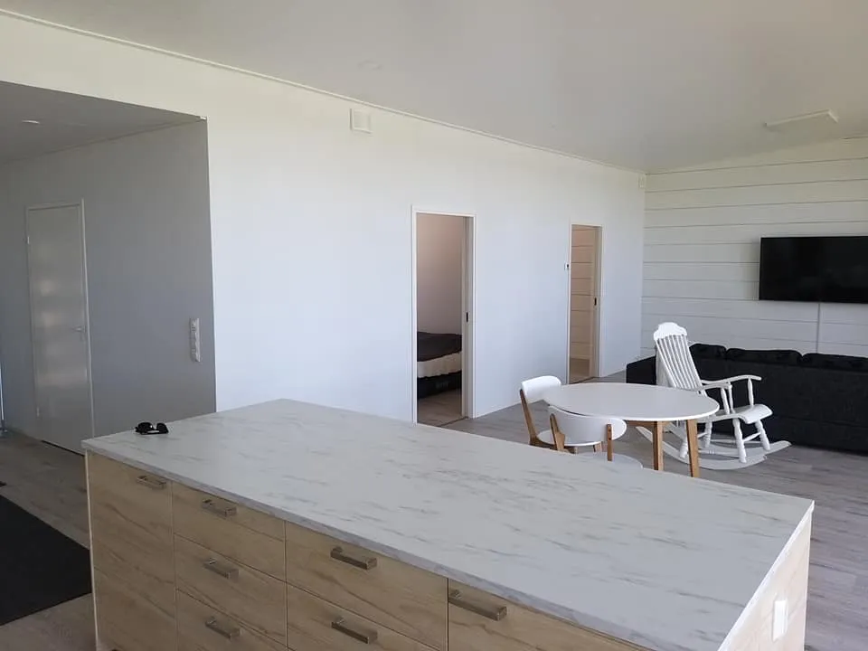 Vaalea sisustus, keittiösaareke, ruokapöytä ja olohuone, jossa on musta sohva ja valkoinen keinutuoli.