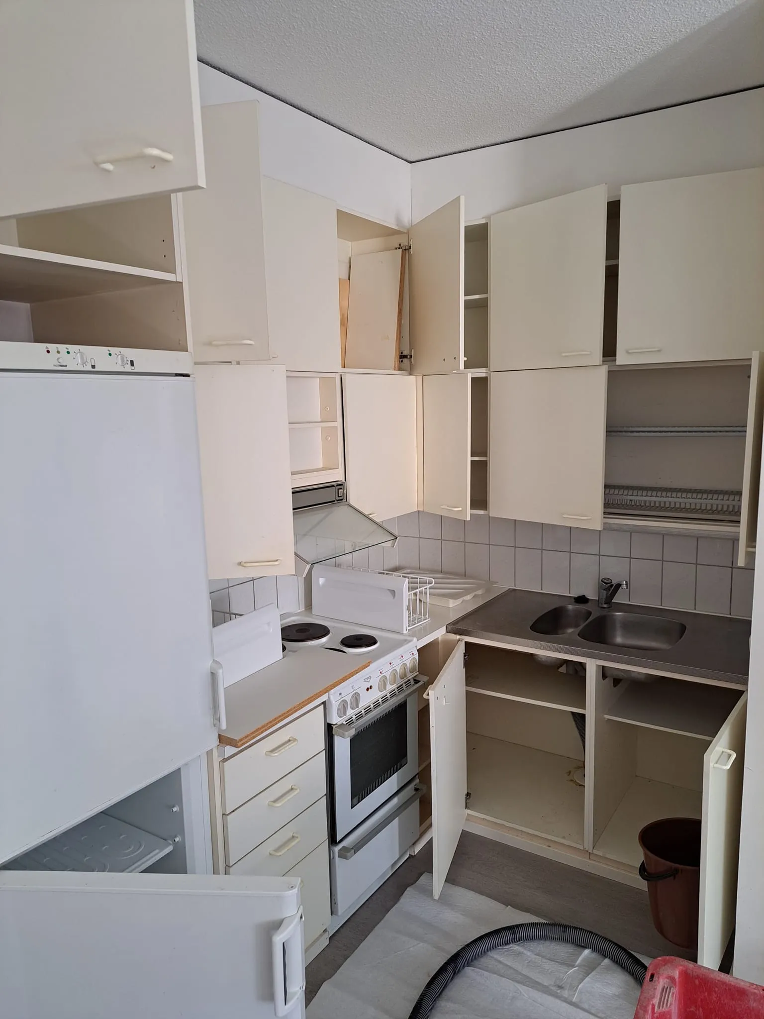 Valkoinen keittiö, jossa avoimet kaapit, pesuallas, liesi ja jääkaappi, valoisa tila.