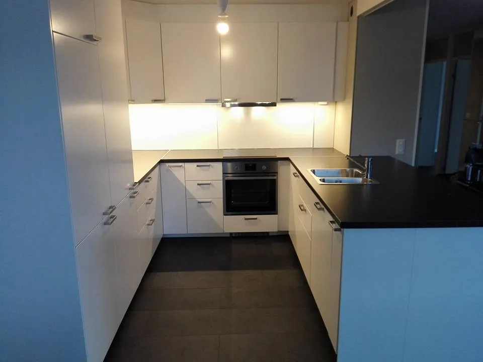 Moderni, valkoinen keittiö, jossa on tumma työtaso, teräksinen pesuallas ja uuni, sekä tummat lattialaatat ja kirkas valaistus.