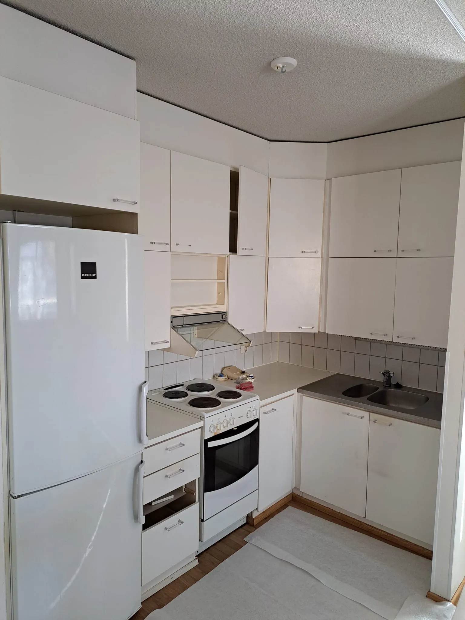 Valkoinen keittiö, jossa on jääkaappi, liesi ja pesuallas, valaistus on kirkas.