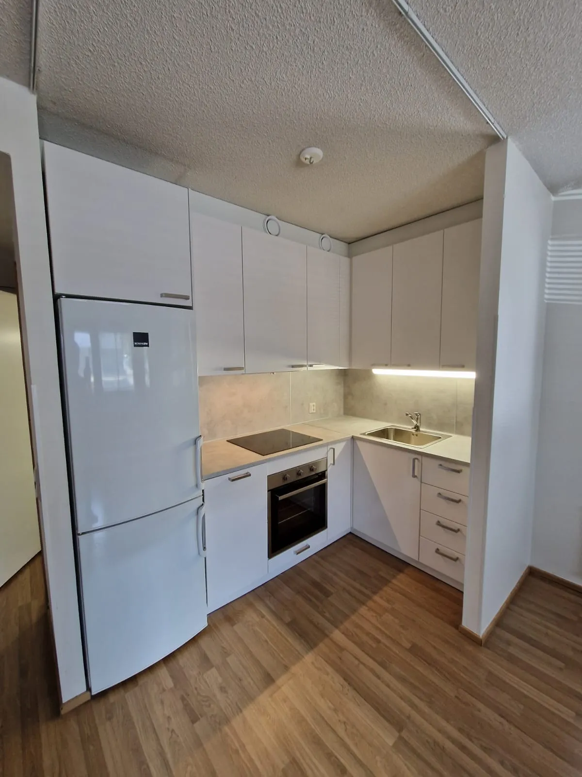 Valkoinen keittiö, jossa jääkaappi, uuni ja laminaattilattia.