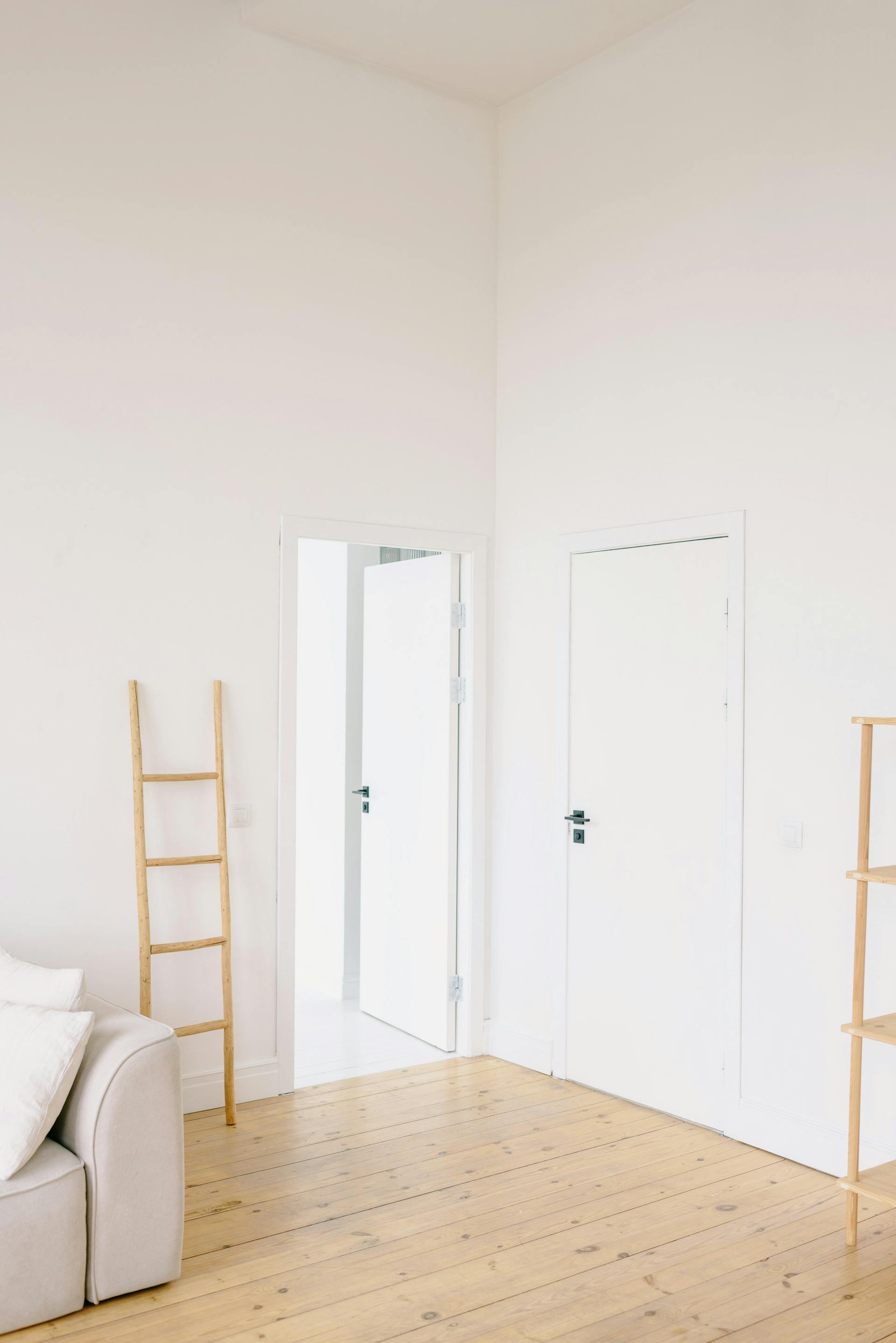 Vaalea huone, jossa puinen tikapuu, avoin ovi ja beige sohva, valoisa ja minimalistinen sisustus.