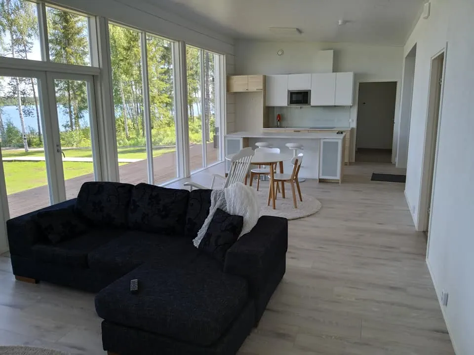 Valoisa ja avara olohuone, jossa on suuri musta kulmasohva, valkoinen keittiö ja suuret ikkunat, joista näkyy järvimaisema.