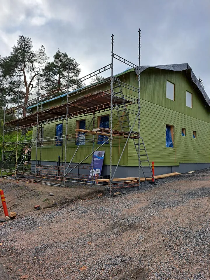 Kaksikerroksinen vihreä puutalo, jota ympäröi rakennustelineet ja jonka ikkunat on suojattu sinisellä muovilla, sijaitsee soratiellä pilvisen taivaan alla.